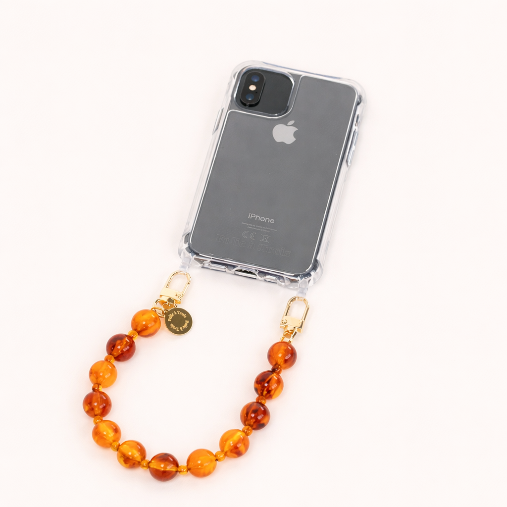 A clear phone case with an amber bead phone strap and a gold Folie à Trois charm, shown on a cream background.