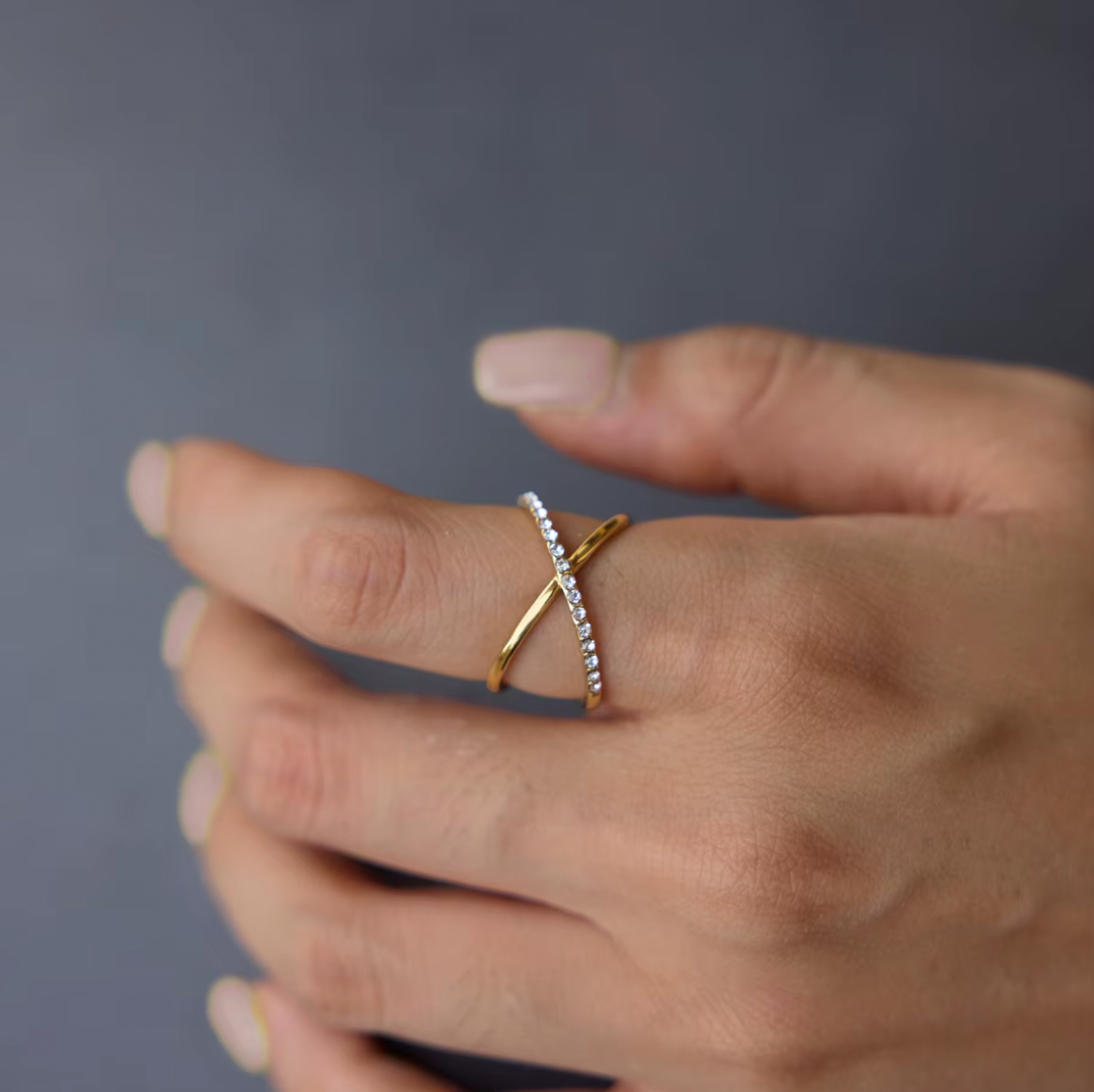 Close-up of the gold Kalimera RING's crossed pavé and plain bands on a model's finger against a dark grey backdrop.