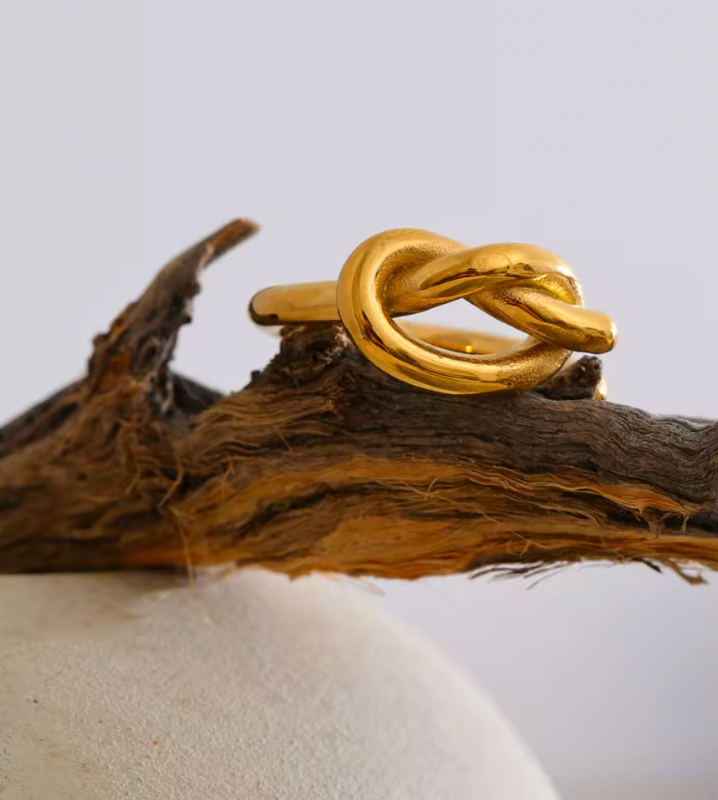 Close-up of the gold Knotted RING's sculpted knot balanced on a weathered driftwood branch against a pale backdrop.