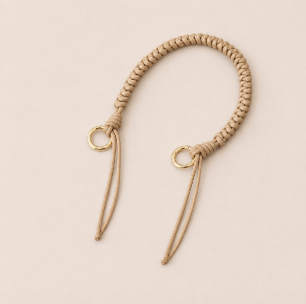 Folie à Trois Layover short wholesale phone strap in beige braided cord with gold rings, shown as a knotted loop with fringe ends on a cream background.