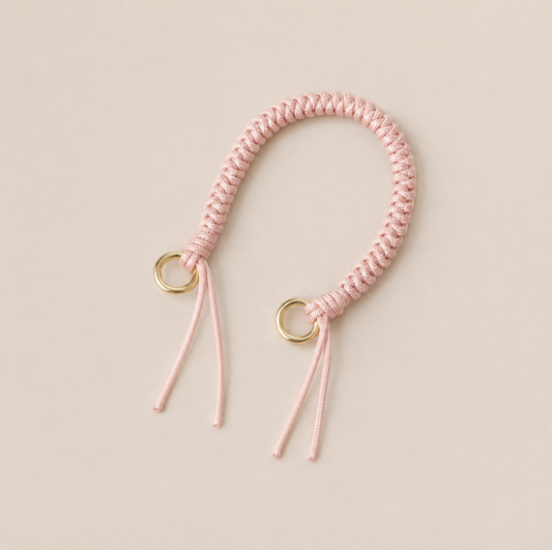 Folie à Trois Layover short wholesale phone strap in pink braided cord with gold rings, shown as a knotted loop with fringe ends on a cream background.