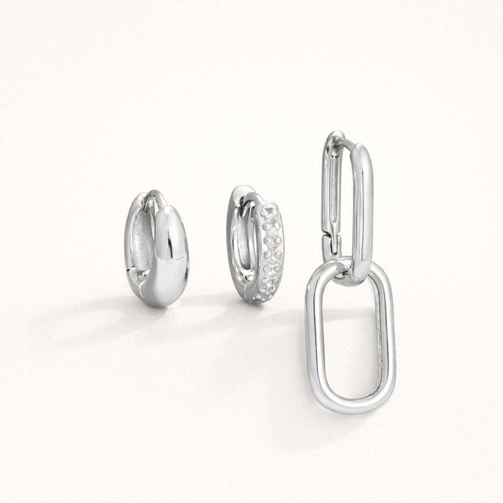 Lea Set in silver tone, a chunky teardrop hoop, a pave hoop, and a paperclip hoop with a large oval charm, laid flat.
