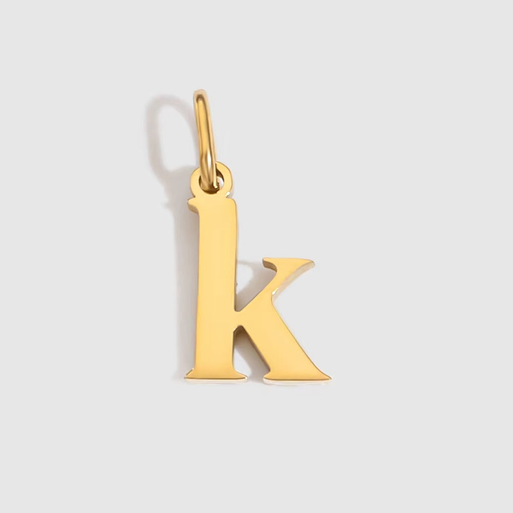 The Letter CHARM in lowercase k, a polished gold pendant in a serif k shape, shown in close-up on a light grey background.