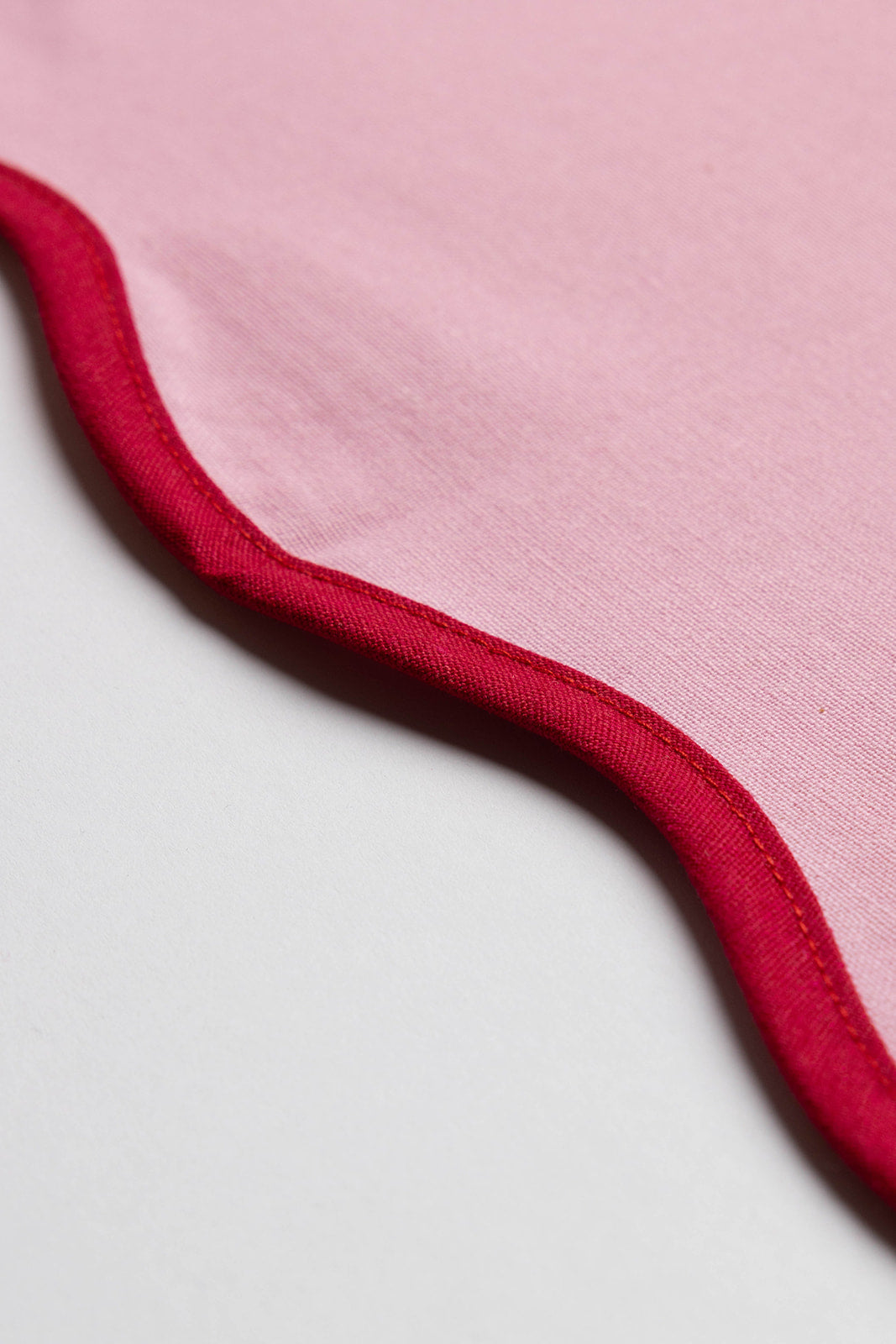 Macro detail of the pink Lilou Scallop napkin's wavy red-trimmed edge against a soft grey background.