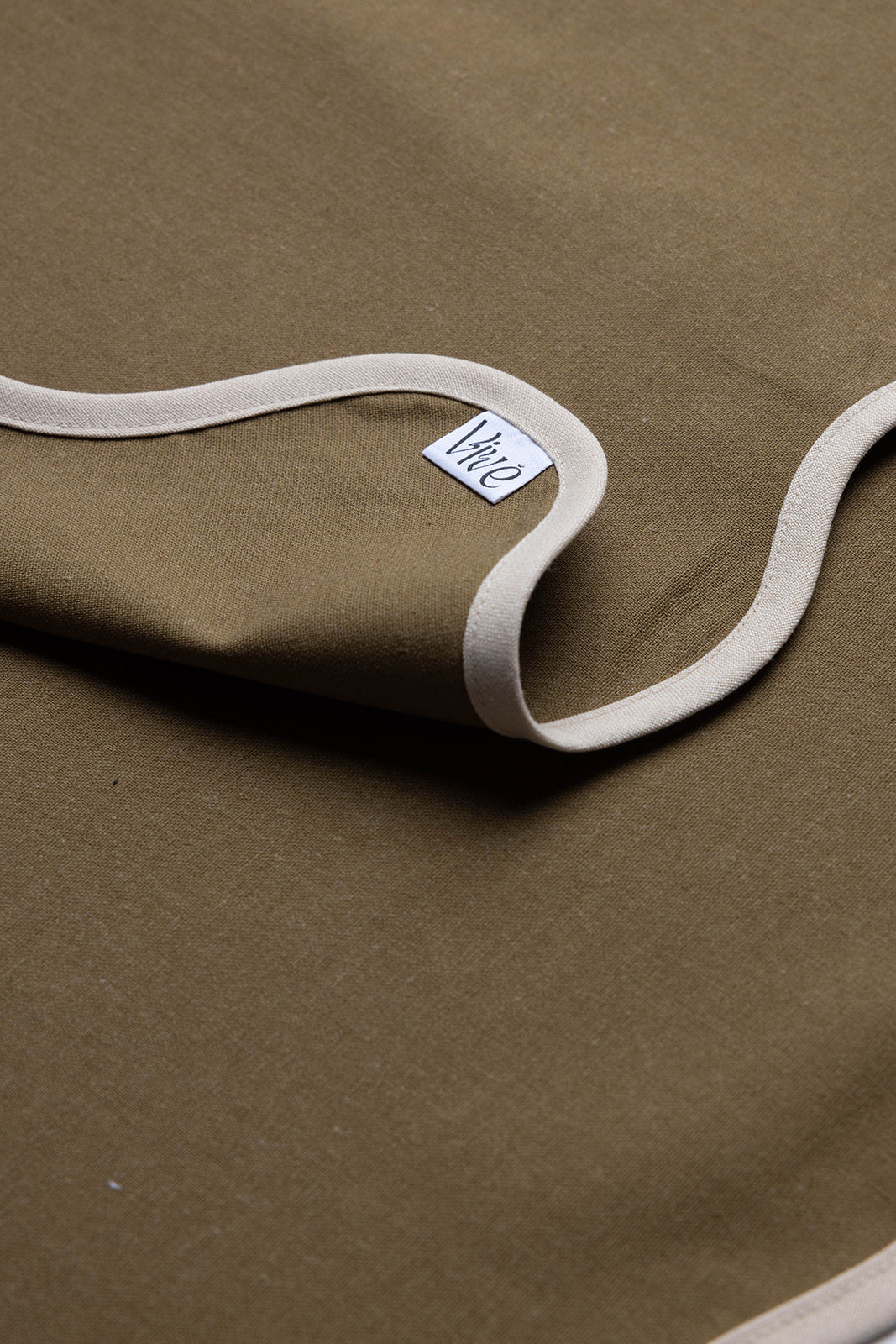 Folded corner of an olive Lilou Scallop napkin reveals the woven Vive brand label against the cream trim.