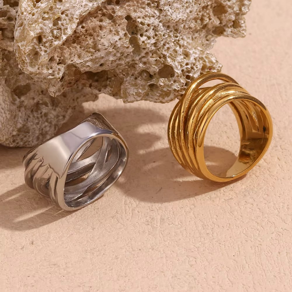 Wide silver band Lizie RING beside its gold wrap-style counterpart, both resting against a textured stone.
