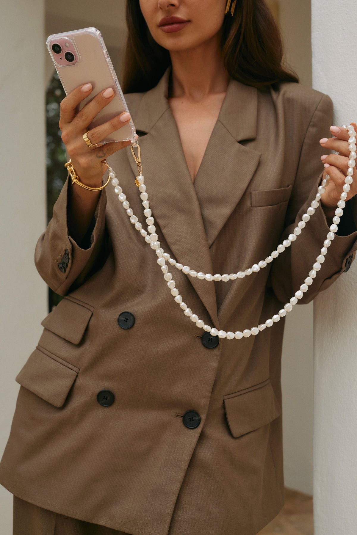 Woman in an olive blazer holding her phone by a long round pearl Long Pearl PHONE STRAP draped from her hand, gold jewelry.