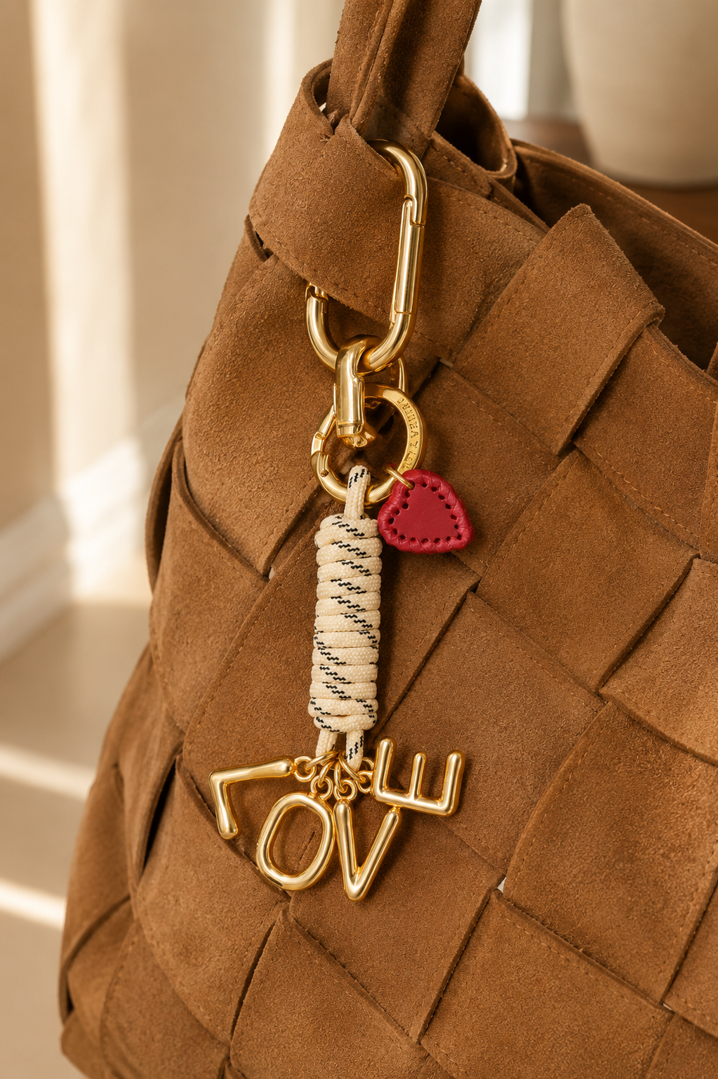 Folie à Trois Love Note BAG CHARM, gold LOVE lettering with a red leather heart charm, clipped to a woven tan suede bag.