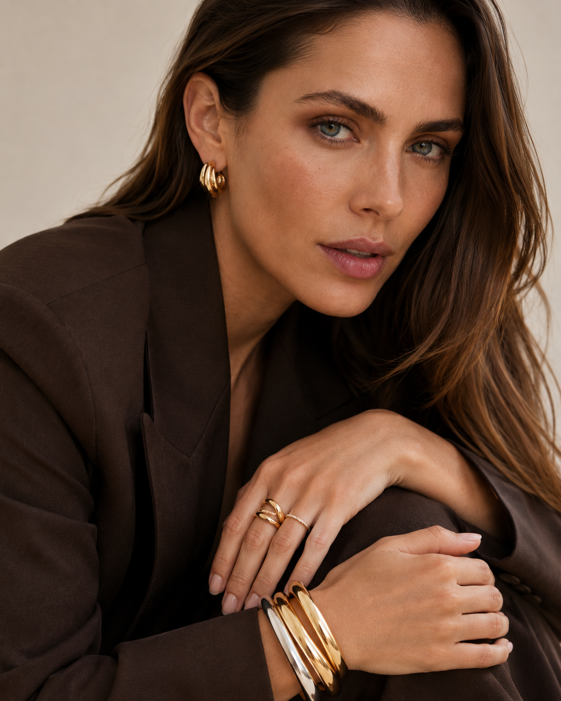 Model in a brown blazer wears the gold Lustro EARRINGS ribbed half-hoop stud, paired with stacked rings and bangles.