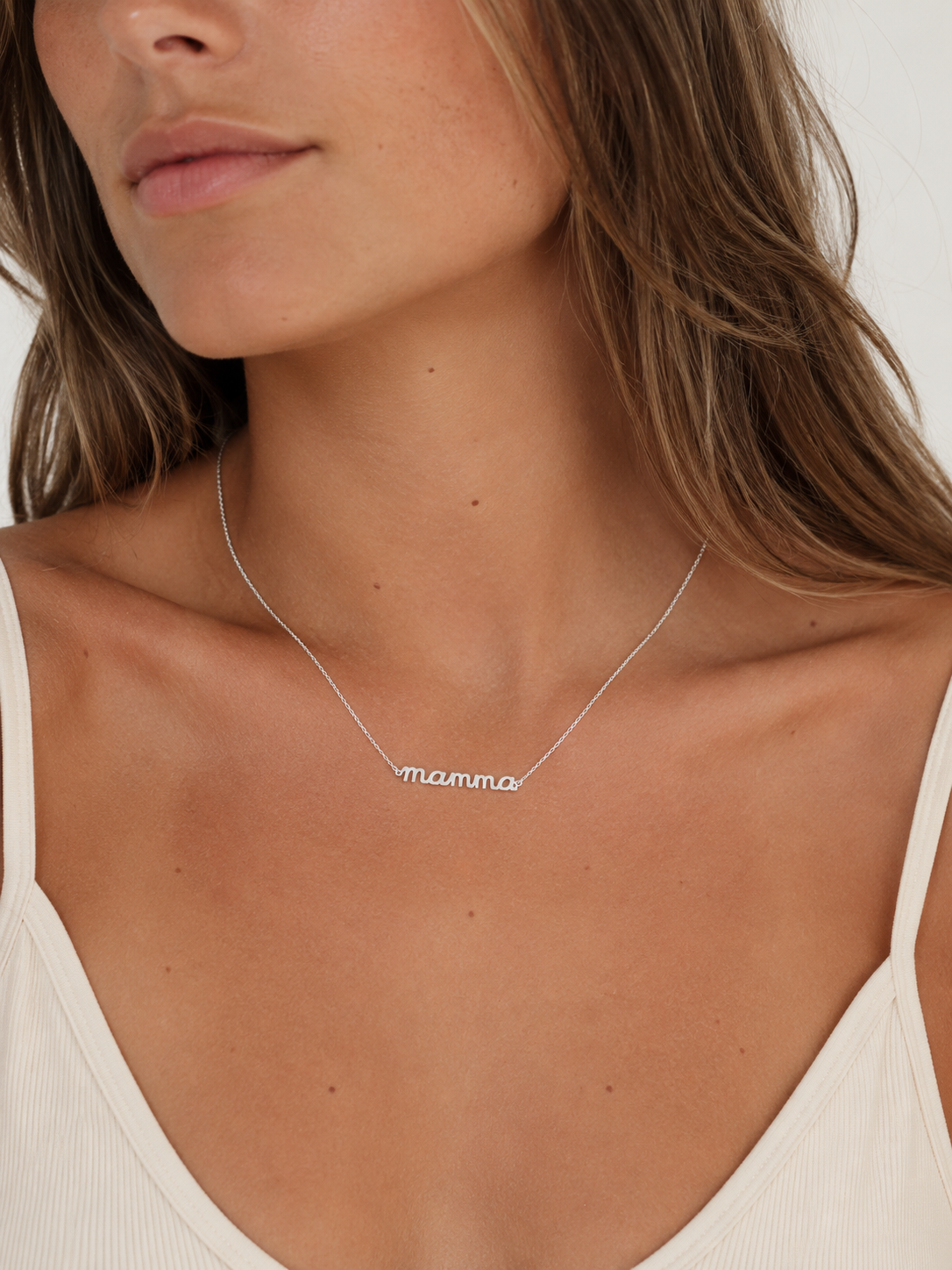 The Mama Big NECKLACE in silver worn at the neckline, a script mamma pendant on a fine chain above a cream tank top.