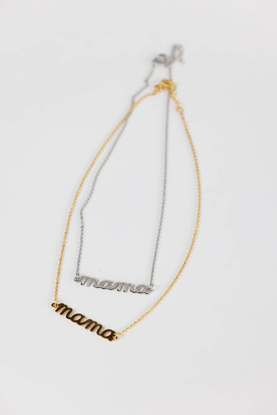 The Mama Big NECKLACE shown in gold and silver together, both script pendants laid side by side on a white surface.