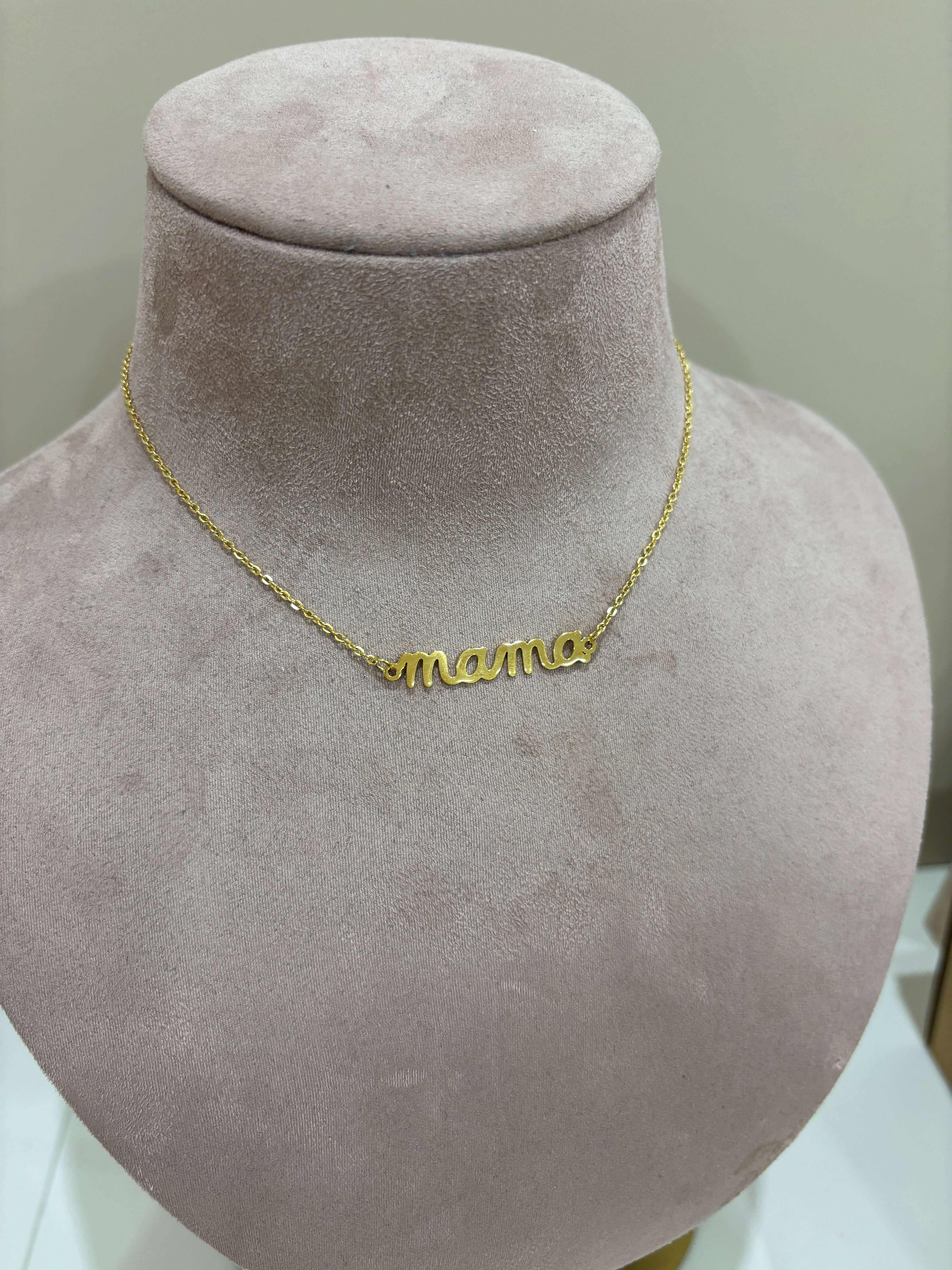 The Mama Big NECKLACE in gold, a script mamma pendant on a fine chain, photographed lying on a white surface.