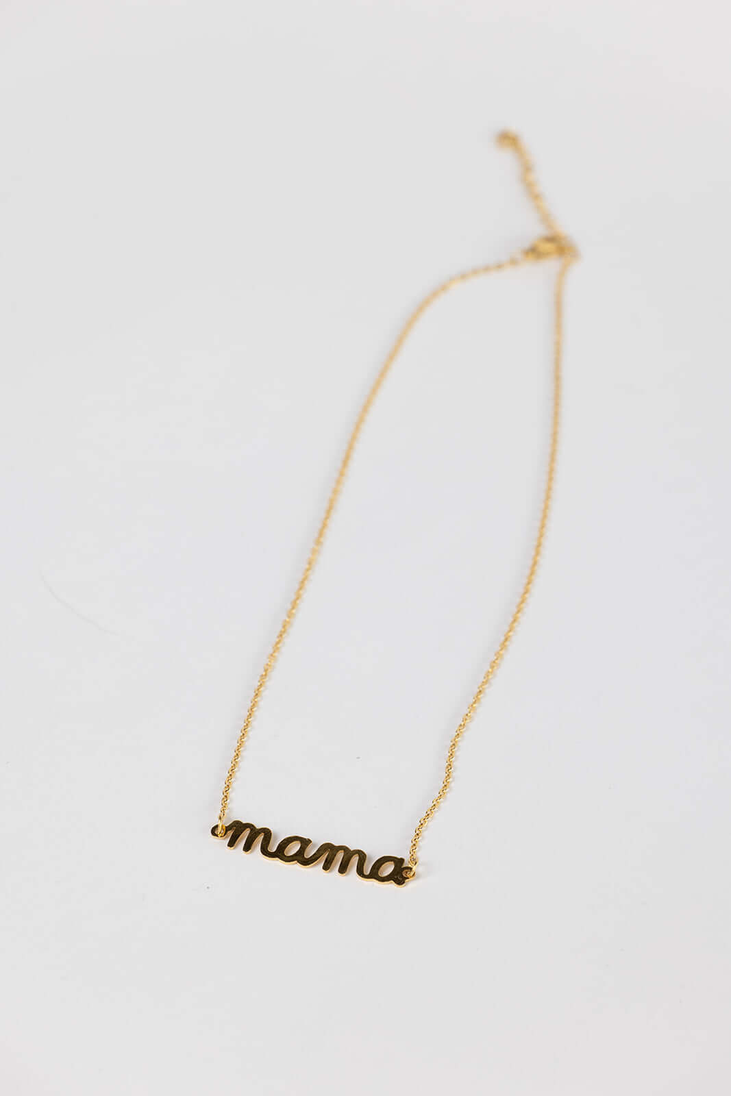 The Mama Big NECKLACE in gold, a script mama pendant on a fine chain, photographed lying on a white surface.