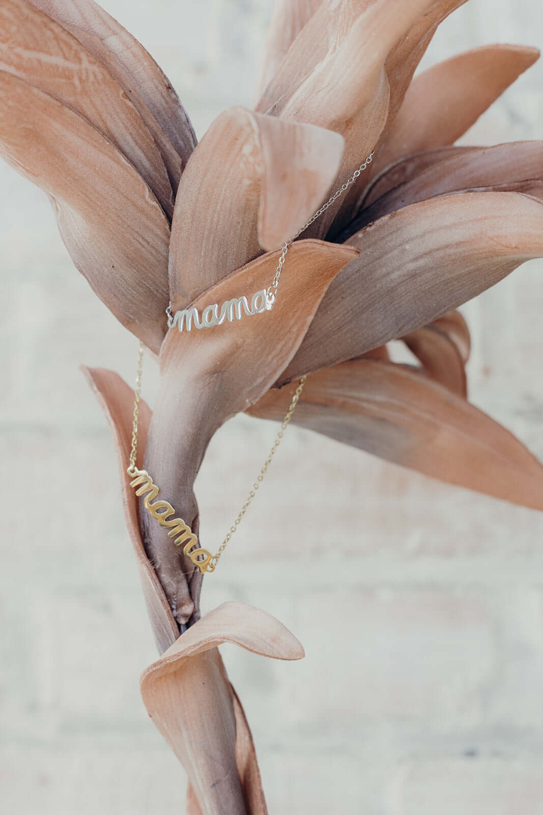 The Mama Big NECKLACE shown in silver and gold, both script mama pendants styled among dried tropical flower petals.