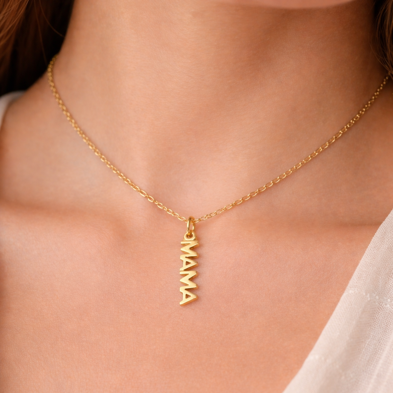 The Mama CHARM worn at the neckline, a gold vertical script MAMA pendant on a fine chain against a cream top.