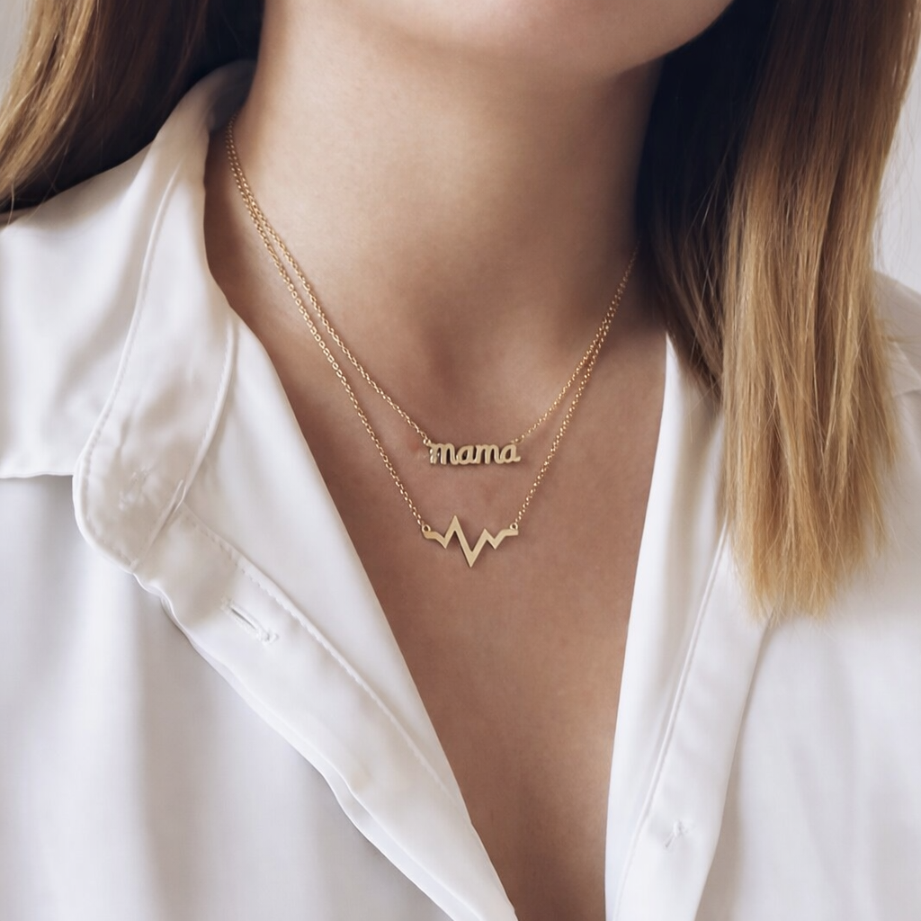The Mama Small NECKLACE in gold layered with a heartbeat pendant necklace, both worn at the neckline over a white shirt.