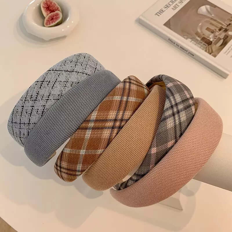 Six different padded headbands - grey tweed, ribbed grey, brown plaid, camel, grey plaid and blush pink - resting together near a dish of figs.