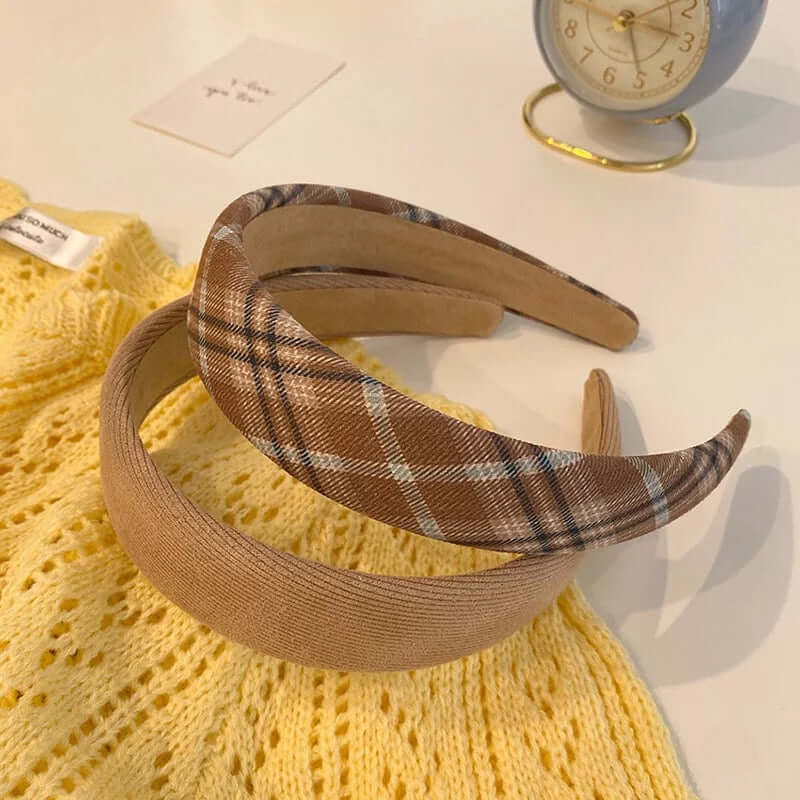 Two padded headbands, one brown and white plaid and one camel ribbed, resting together on a yellow knit sweater.