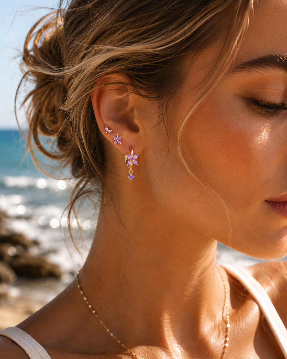 A model wears the three-piece Misha SET gold earrings in purple stacked along her ear, including a flower hoop with a drop charm.