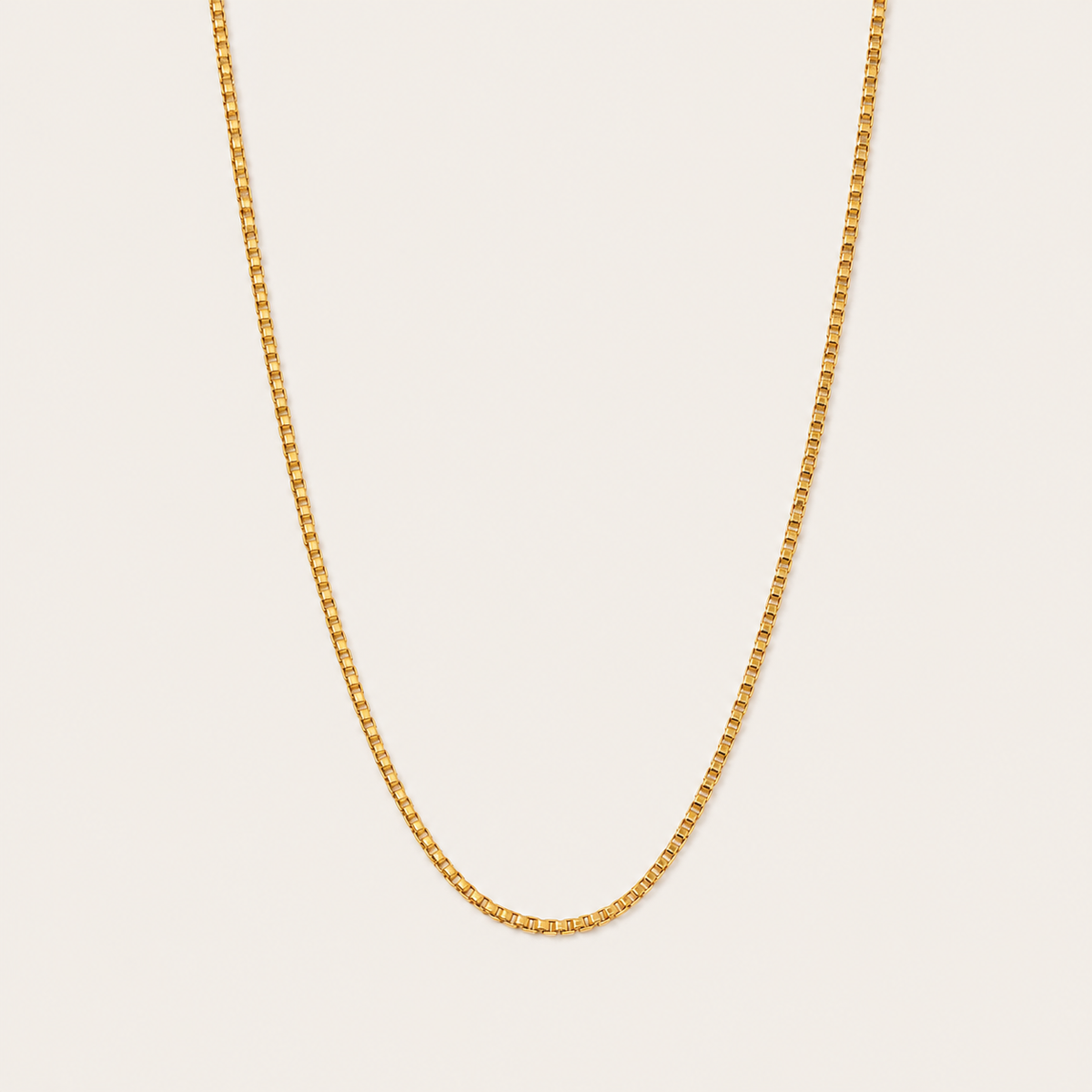Gold box-chain necklace with a fine square-link design, the Nara NECKLACE, shown in a close-up studio shot on a beige background.