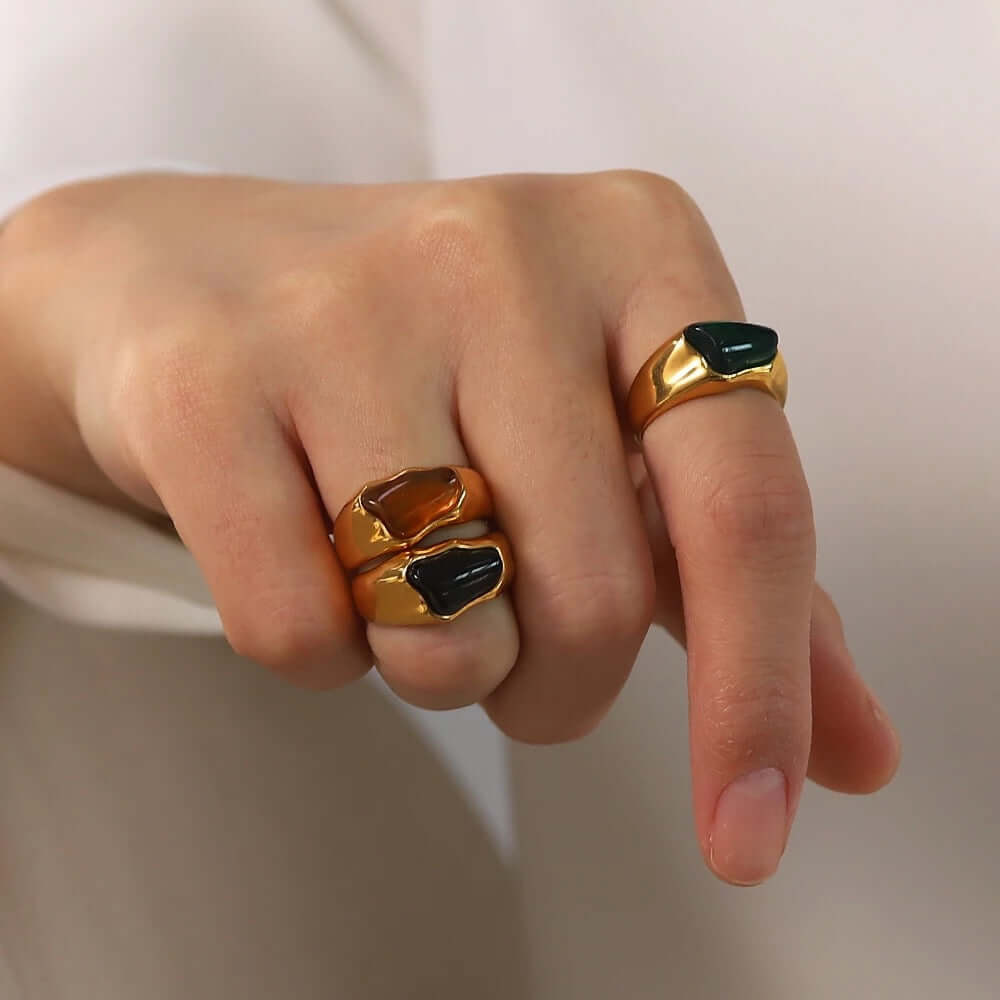 Close-up of a hand wearing two stacked gold signet-style New Fashion RING styles, set with amber and black cabochon stones.