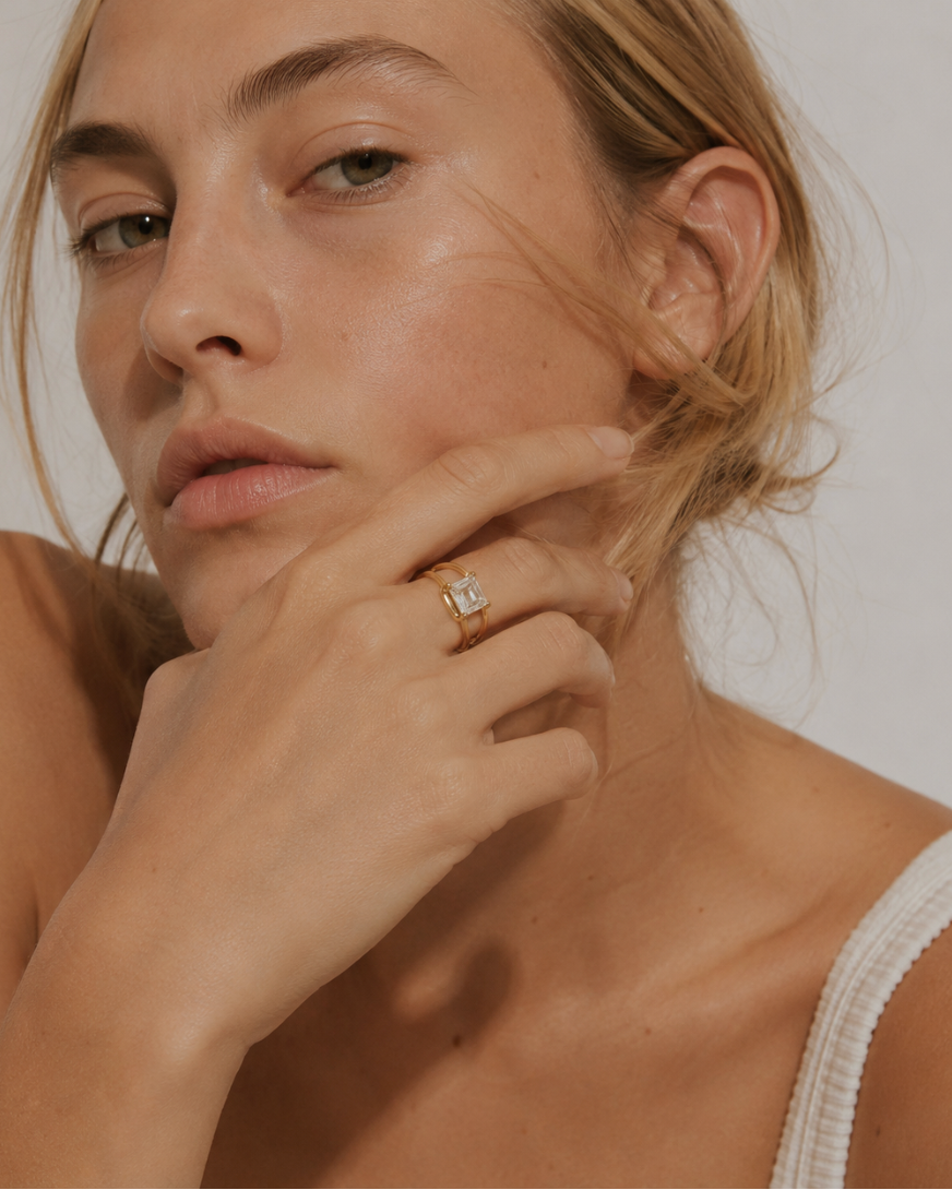 Model wears a gold Ovale RING with an emerald-cut white stone, hand raised to the chin — Folie à Trois