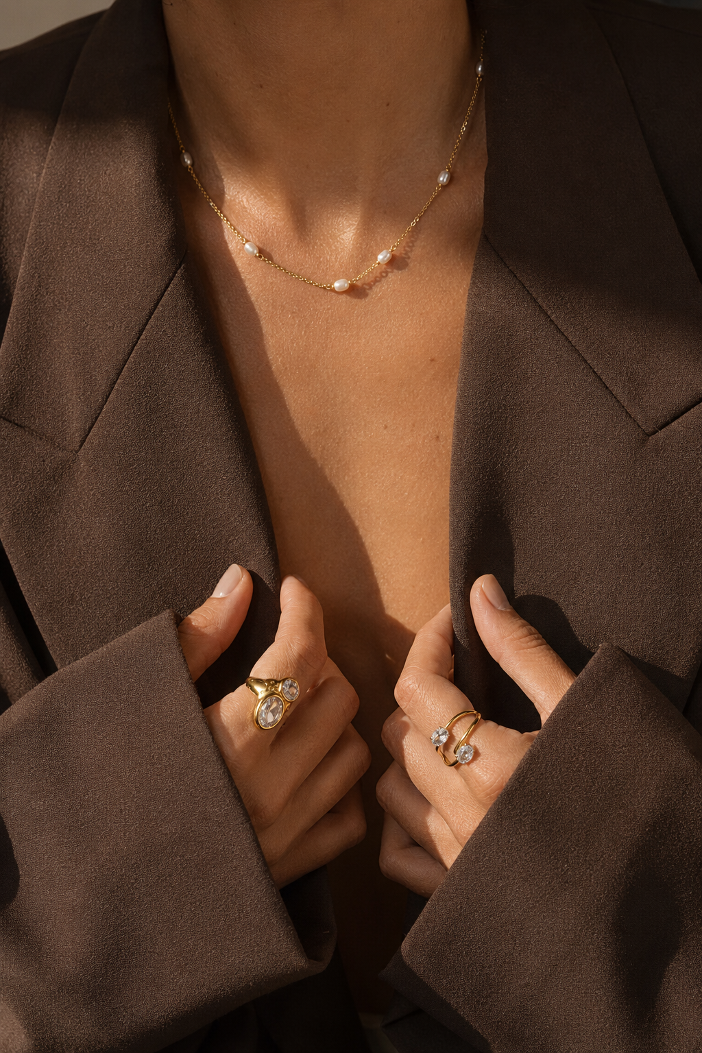Close-up of a person wearing gold jewelry, including a delicate gold necklace with small pearl-like beads and multiple gold rings with clear stones, styled with a dark brown blazer.