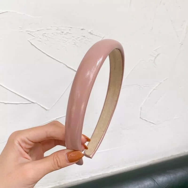 A single glossy mauve pink patent-look padded headband held up by hand against a textured white wall.