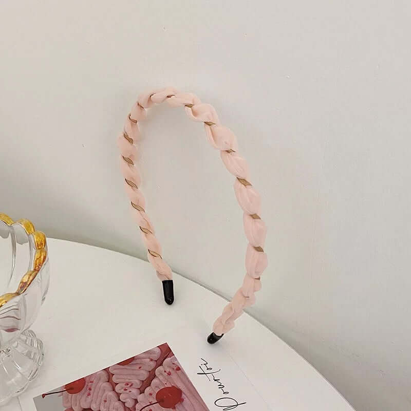 A single pale pink braided twisted headband with black tips styled upright near a glass dish and photo card.