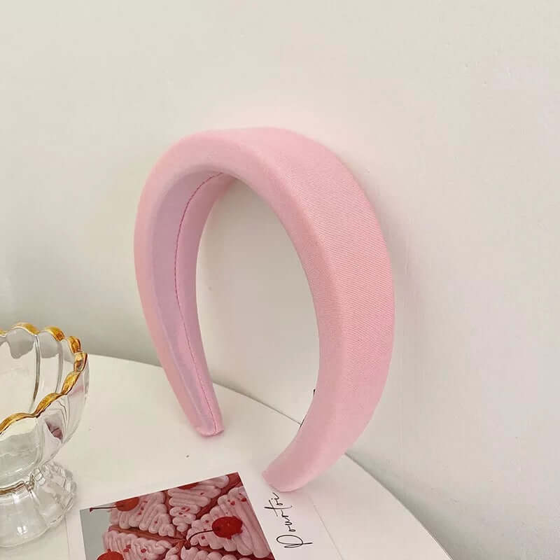 A single pink grosgrain-ribbed padded headband styled upright near a glass dish and small photo card.