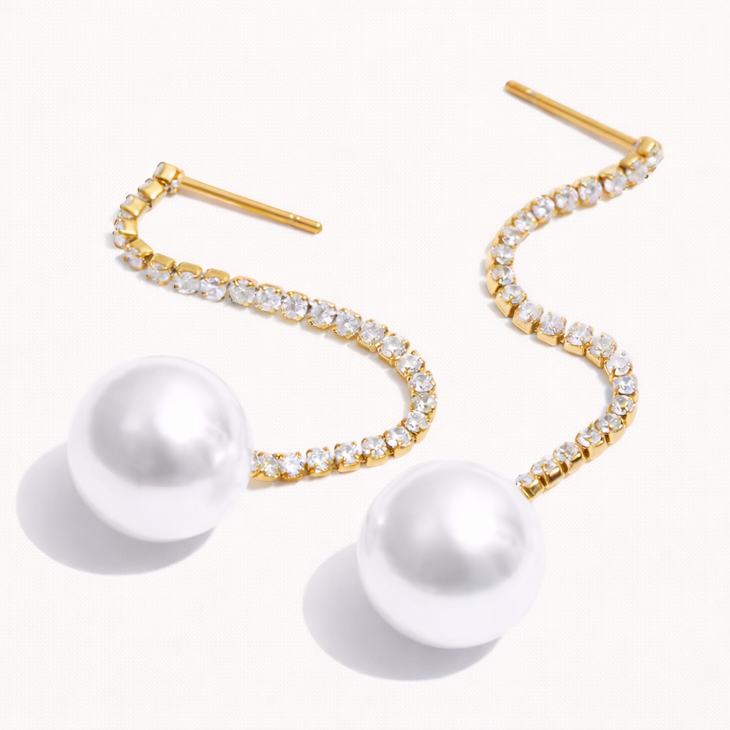 Rhea Earrings featuring a curved gold chain set with sparkling stones that spirals down to a lustrous white pearl drop, shown as a pair.