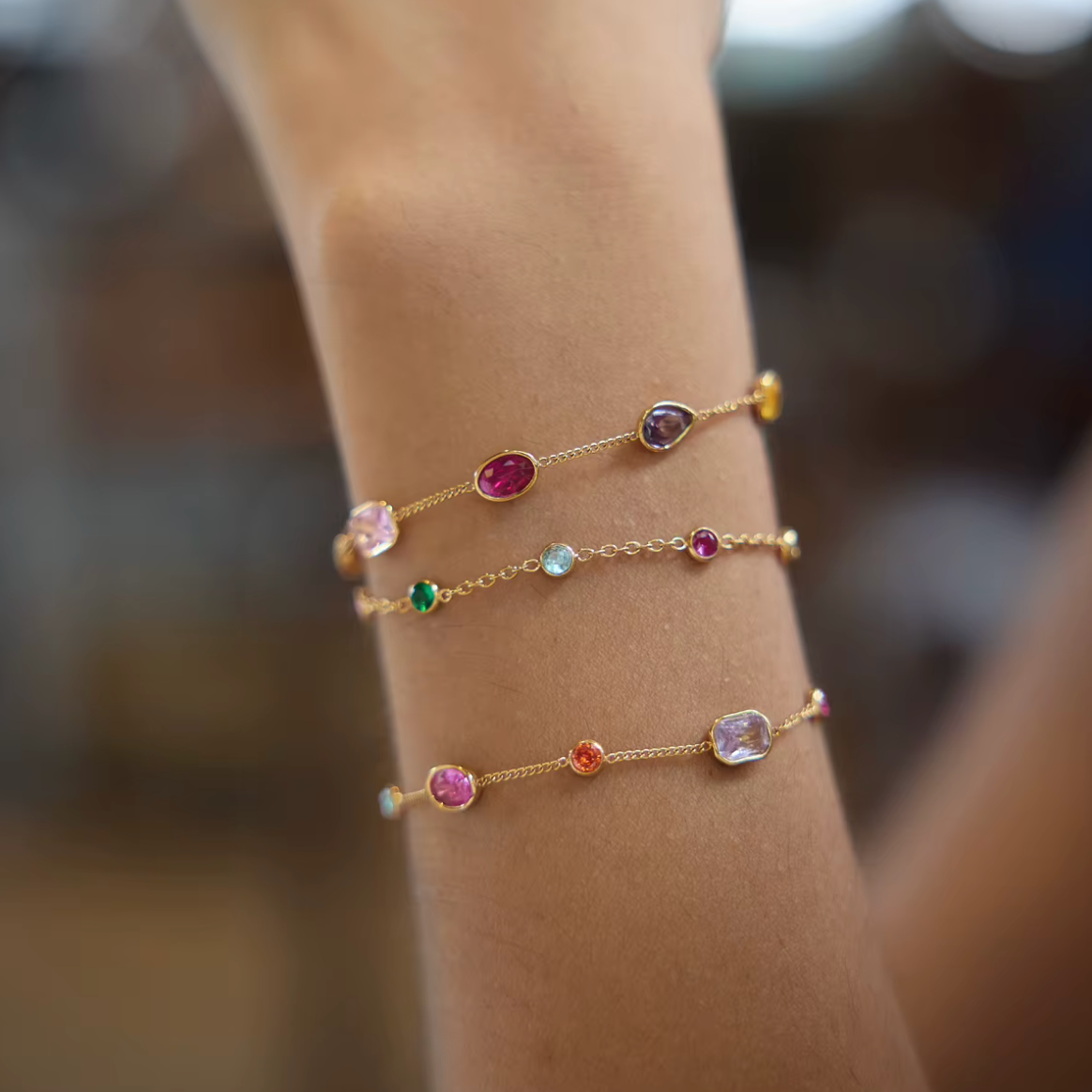 Three gold Riva BRACELETs stacked on a raised wrist, set with pink, purple, green and yellow bezel gemstones on fine chains.