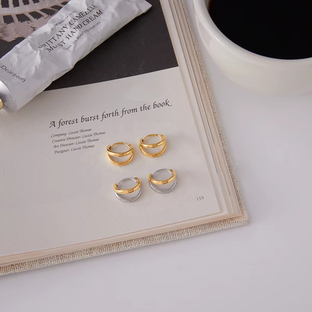 Roadtrip Earrings styled on an open book page beside a hand cream tube and a cup of coffee, in gold and two-tone finishes.