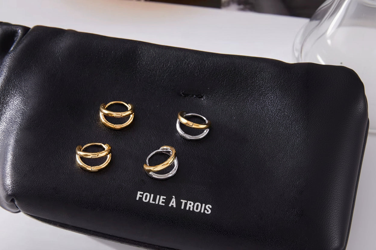 Roadtrip Earrings in gold and mixed two-tone finishes resting on a black pouch printed with the Folie a Trois logo.