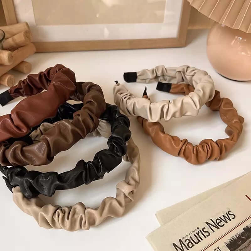 Five ruched faux leather Romy HAIRBAND colorways in beige, black, chestnut and cream, arranged together beside a folded newspaper prop.