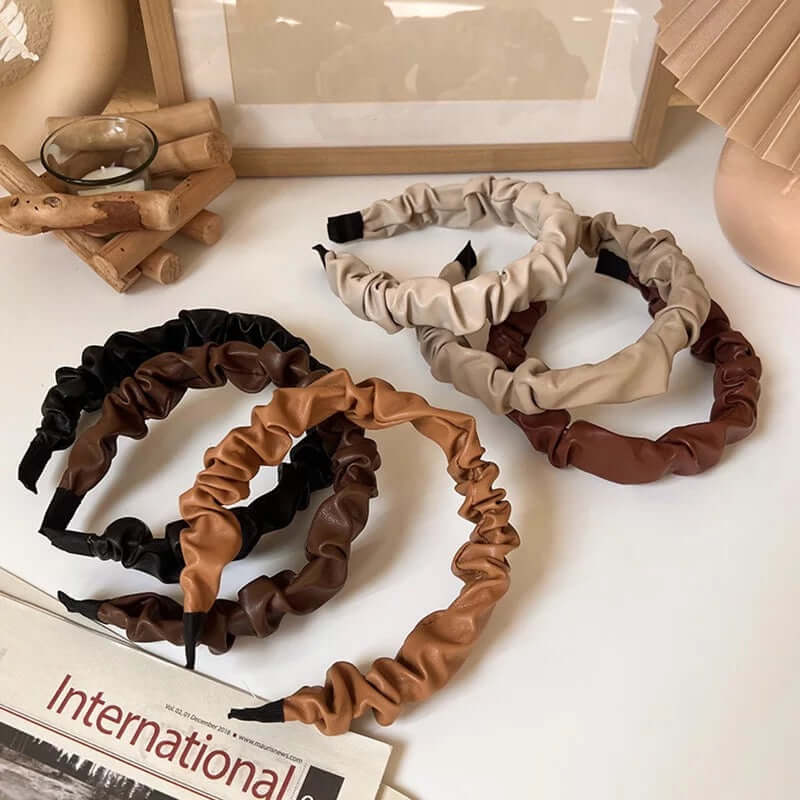 Four Romy HAIRBAND ruched faux leather headbands in black, chestnut, camel and cream, styled together beside an International newspaper.