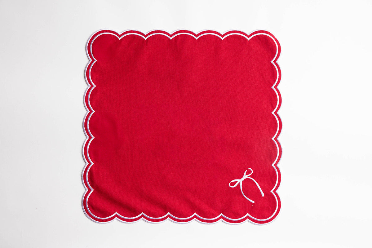 Flat lay of a single red square napkin with white scalloped edging and an embroidered bow, the Rosie Scallop napkin design.