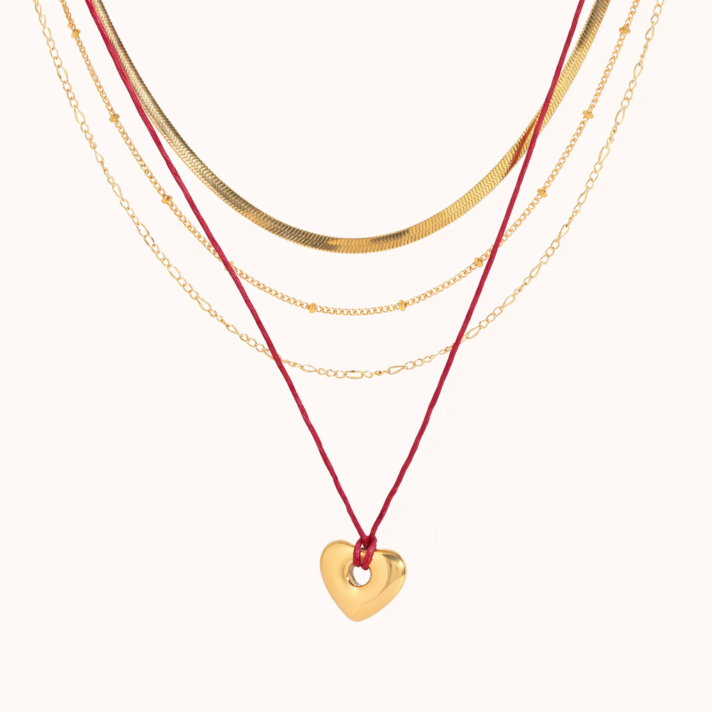 The Rowie NECKLACE, three layered gold chains with a red cord and open gold heart charm, shown on a cream background.