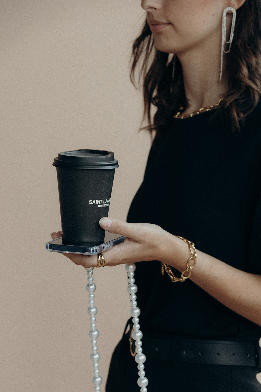 Model holding a phone with the Folie à Trois Sabine Pearl phone chain draped from it, paired with a gold chain bracelet and a coffee cup.
