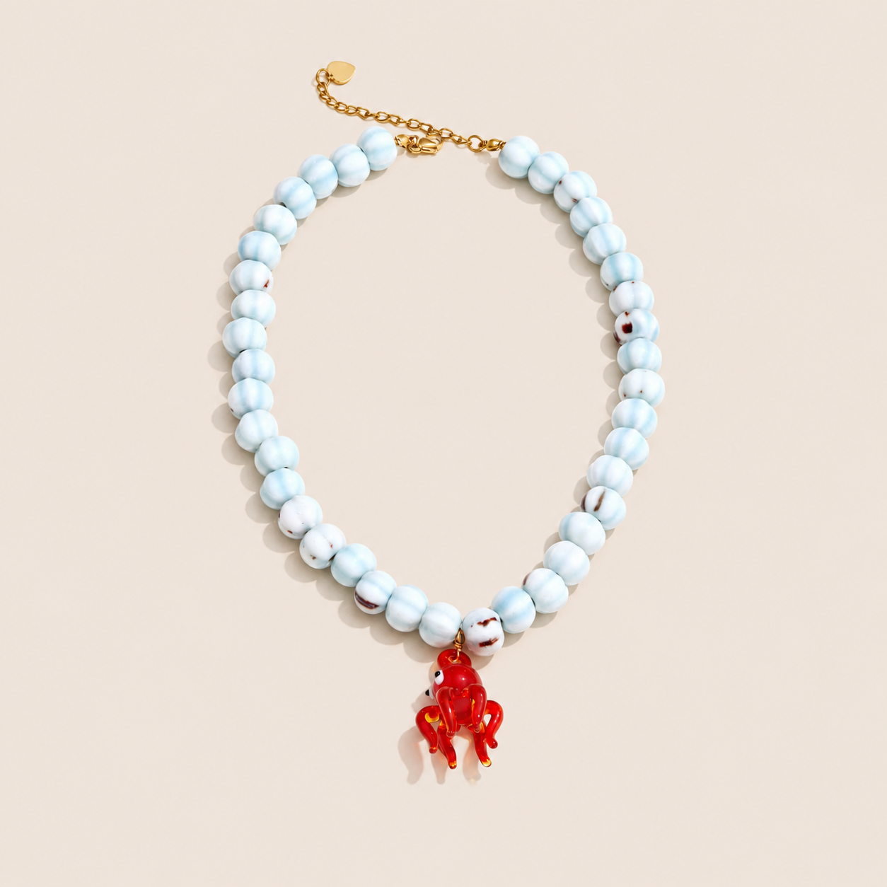 Salt & Sun NECKLACE with a chunky beaded pale blue chain and a red glass octopus charm, laid flat on a beige background.