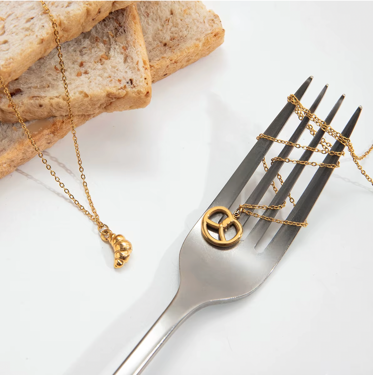 The Salty Sweet NECKLACE and its matching croissant charm necklace looped around the tines of a fork beside sliced bread.