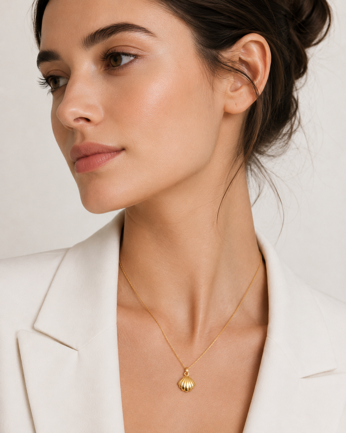 The Shell CHARM worn as a pendant on a fine gold chain necklace, resting at the neckline over a cream blazer.