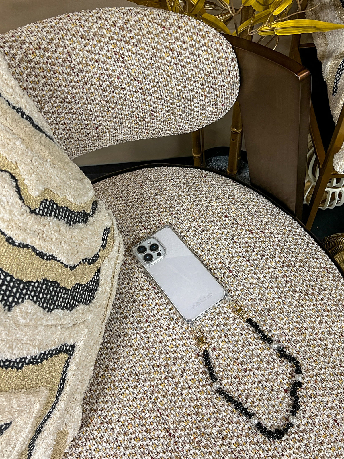 Black beaded Shell Whisper SHORT PHONE STRAP attached to a phone, resting on a tweed chair with gold decor in the background.