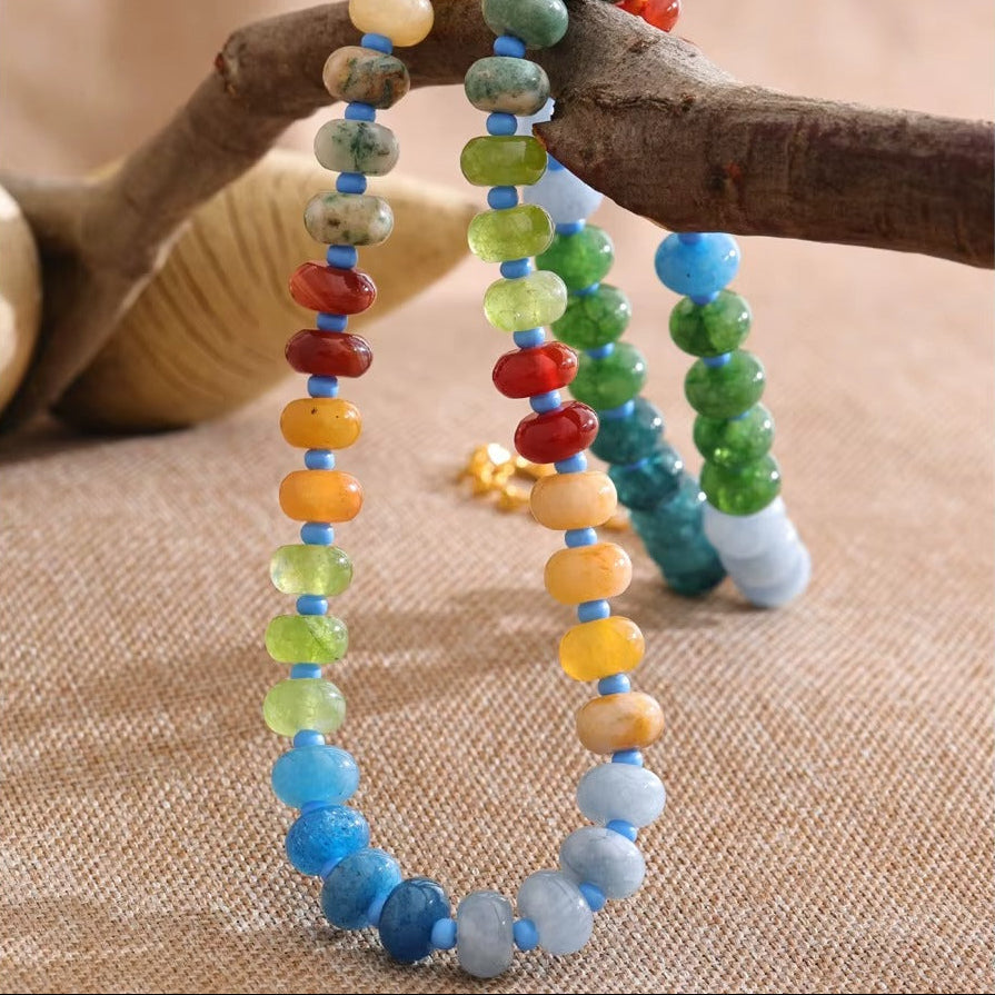 The Sicily NECKLACE beads shown in close detail, draped over a driftwood branch beside a shell, highlighting each stone's color.