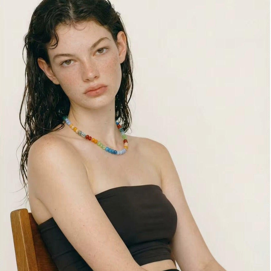 The Sicily NECKLACE worn over a dark strapless top, the model seated on a wooden chair and gazing to the side.