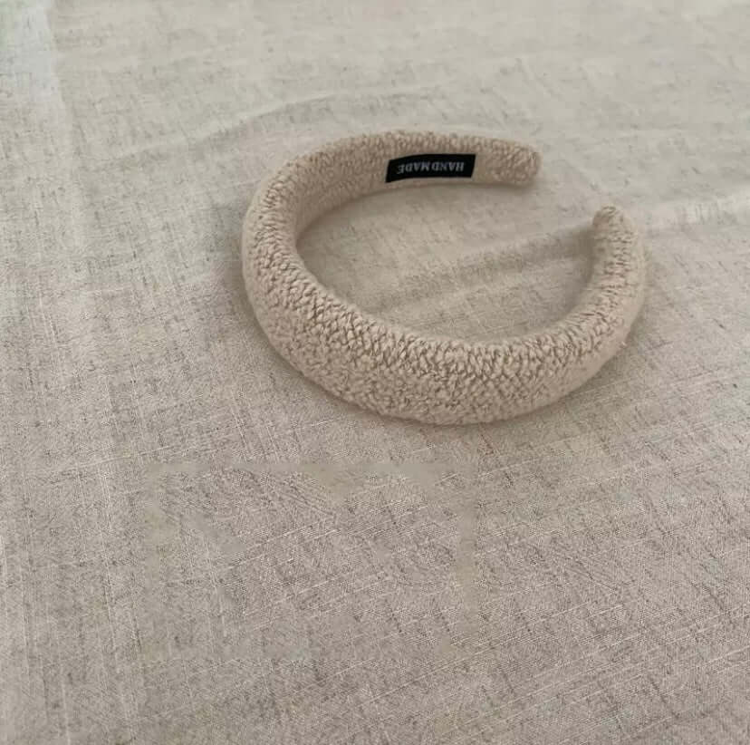 A single cream oatmeal boucle padded Skylar HAIRBAND with a handmade label tag, laid on linen fabric.