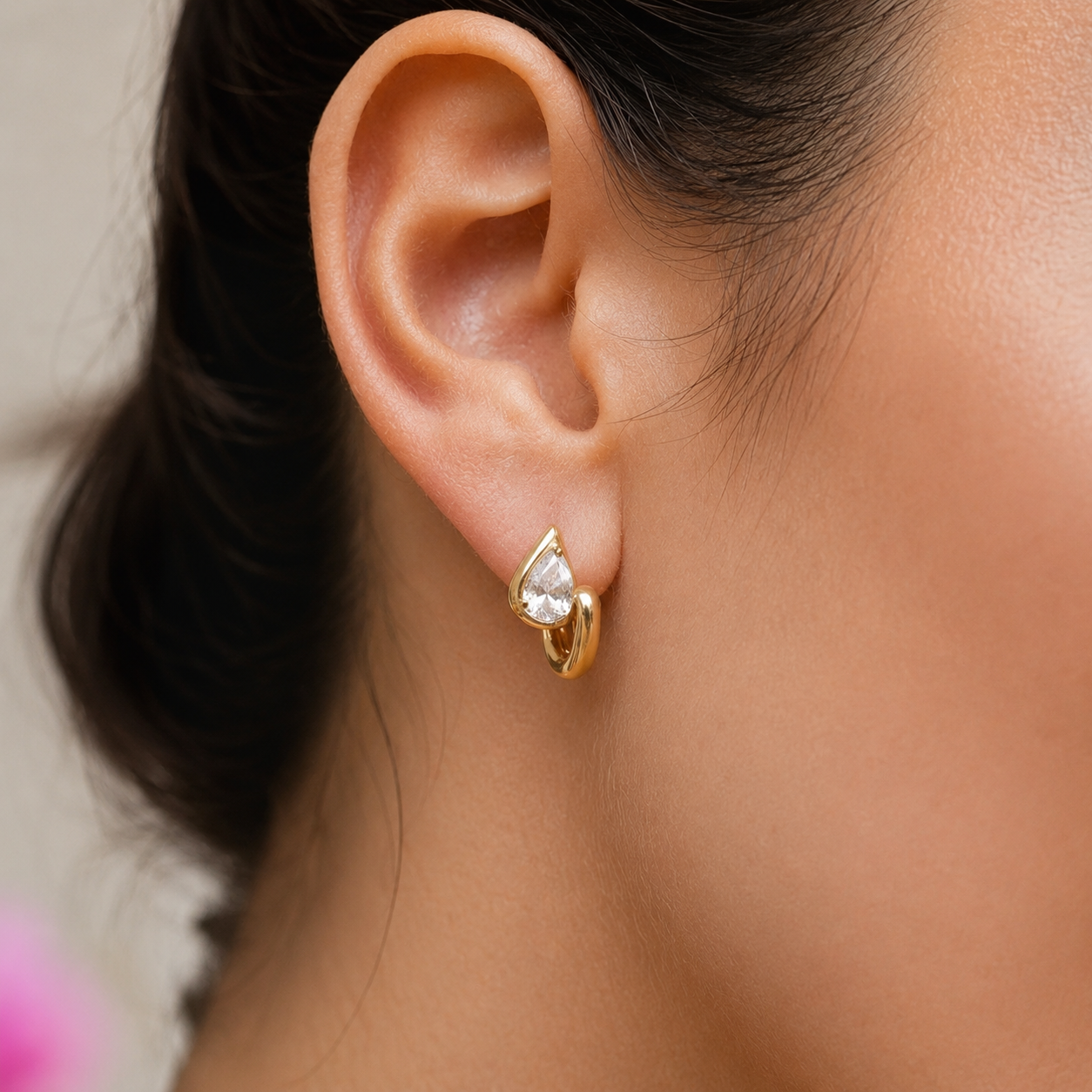 Close-up of the Folie à Trois Sombra EARRINGS, a gold heart-shaped huggie hoop with a pear-cut zirconia stone, worn on a swept-back ear.