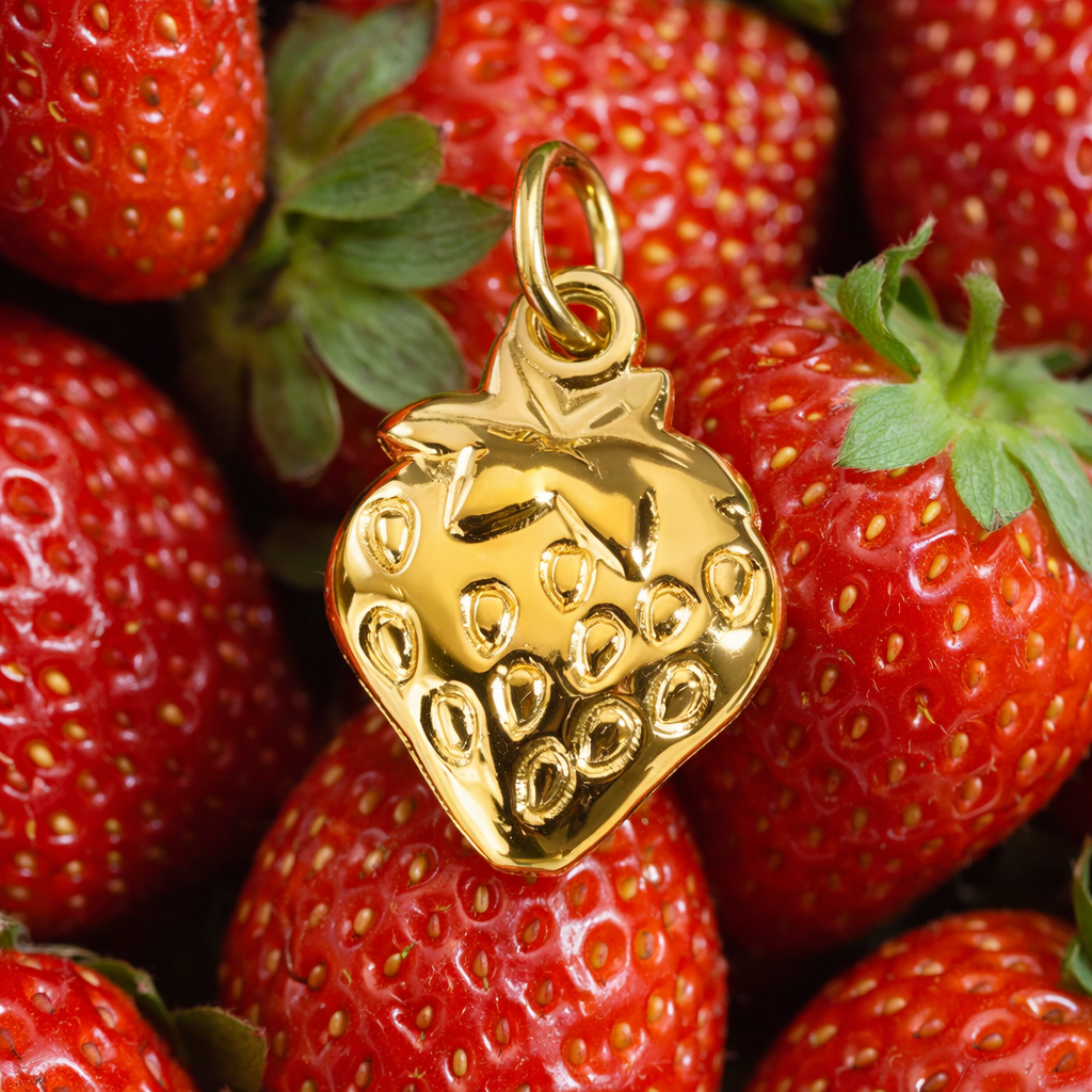 The Strawberry CHARM in gold with textured seed detailing, styled among real strawberries for a playful close-up shot.