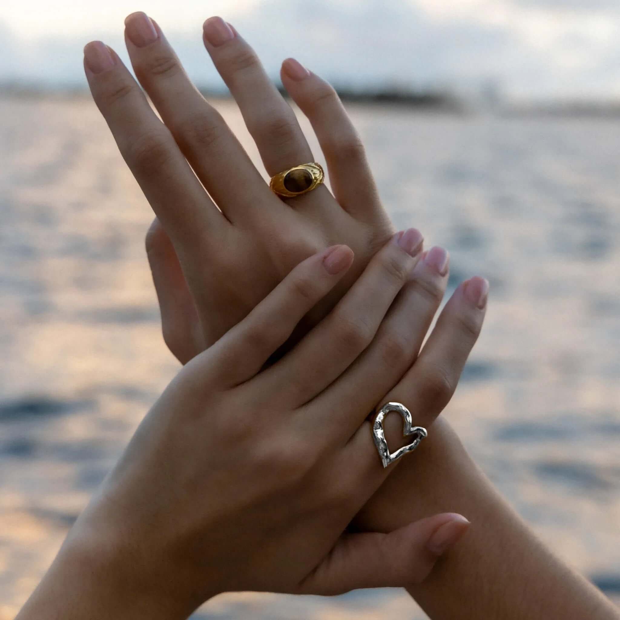 Close-up of clasped hands near the ocean, one wearing the open silver heart-shaped Street Side RING, the other a gold dome ring.