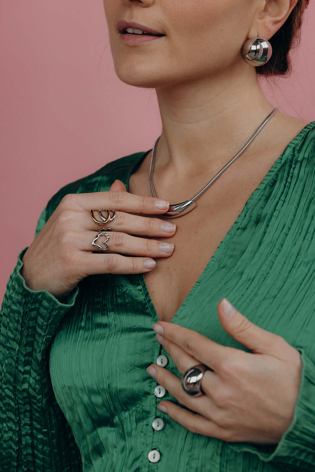 Close-up of a hand near the neck wearing the silver heart-shaped Street Side RING stacked with other rings, pink backdrop.
